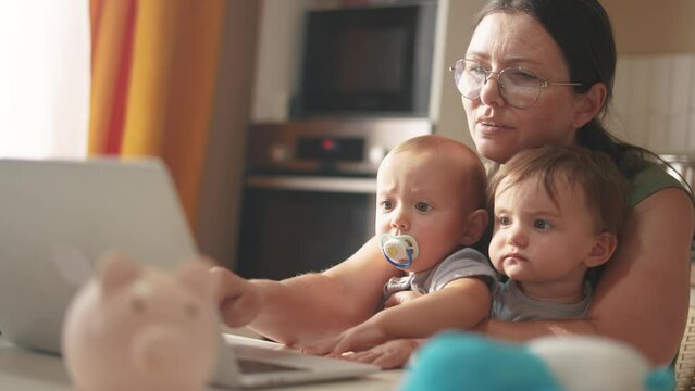 mother works remotely from home with two babies in her arms. pandemic remote work concept. mom tries to work at home in a fun kitchen, small children interfere with business sitting in her arms