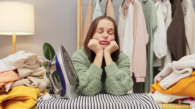 Upset displeased brown haired young woman wearing knitted shirt ironing clothes in her wardrobe at home being lazy to iron looking at camera with sad bored face