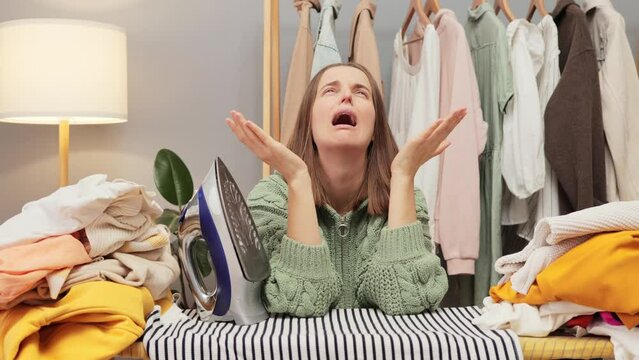 Despair brown haired Caucasian young woman housewife wearing knitted shirt being lazy ironing clothes crying needs rest posing in her wardrobe at home