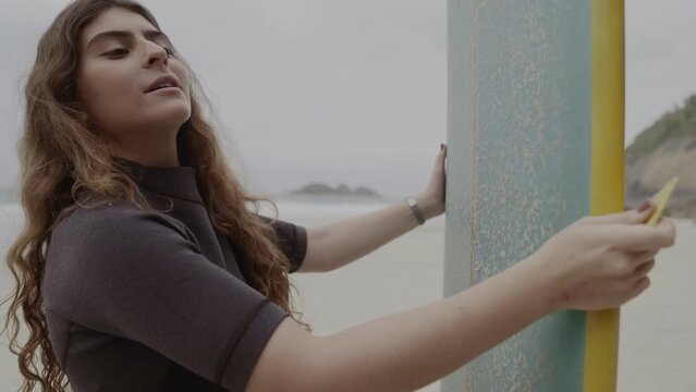 Jovem na praia, concentrada, aplicando parafina na prancha de surf. Cinematico 4k.