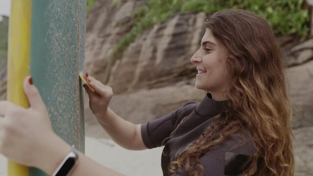 Smiling young woman on the beach applying paraffin to her surfboard. Cinematic 4k.