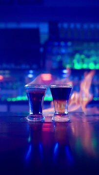 Vertical Video, Two Glass of Alcoholic Drink on a Bar Counter Close-up. Bartender Prepares a Drink by Setting a Glass of Cocktail on Fire in a Nightclub. Bar in Neon Blue Lighting. Slow Motion.