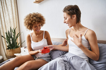 happy african american woman holding red present near her girlfriend in pajamas on valentines day