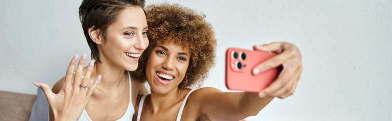 happy and engaged interracial lesbian couple taking selfie on smartphone at home, banner