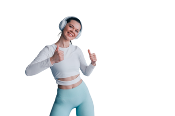 Brunette hispanic young woman in headphones, sportswear stands against grey wall shows thumb up gesture toothy smiles satisfied by great exercise against transparent background. Healthy lifestyle
