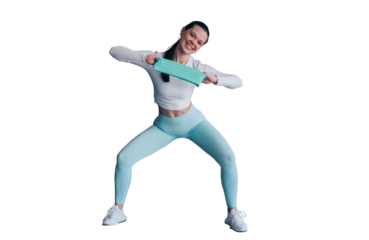 Brunette young woman in sportswear stretching rubber tape at fitness club looks at camera happily. Fit caucasian girl training against transparent background. Fitness, sport and healthy people