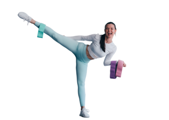 Brunette fit girl in sportswear makes high kick looks at camera holds training rubber tapes happy to be healthy. American young woman against grey wall rising leg up against transparent background