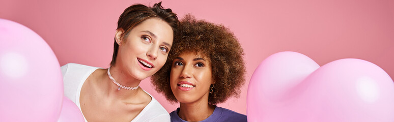 dreamy multicultural lesbian couple looking away near heart shaped balloons, Valentines day banner
