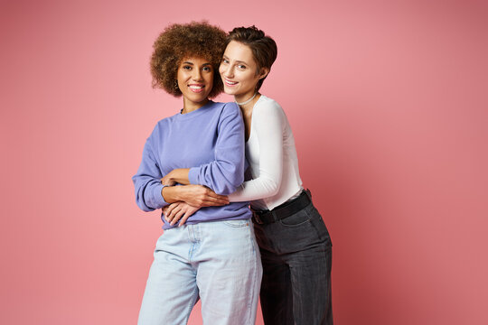 cheerful lesbian woman embracing african american girlfriend in casual clothes on pink background