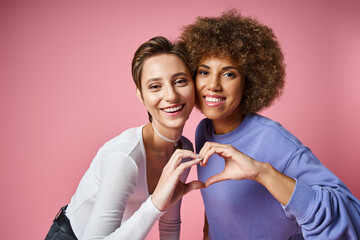 happy multicultural lgbtq couple showing heart sign with hands on pink background, love