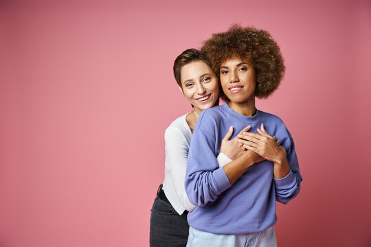 cheerful woman hugging her african american girlfriend on pink background, lgbtq couple love