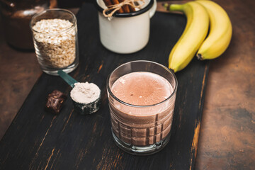 Chocolate milkshake smoothie, protein drink in a glass on dark wooden board with bananas, protein powder in measuring spoon and chocolate pieces