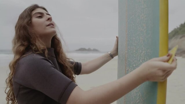 Jovem na praia, concentrada, aplicando parafina na prancha de surf. Cinematico 4k.