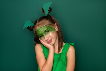 An attractive teenage girl with makeup in the guise of a dragon looks at the camera with a smile. Headdress in the shape of green wings