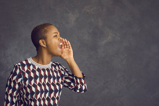 Emotional black woman stands on a gray background and shouts loudly holding his hand to his mouth. Woman with a short haircut making a loud announcement while standing near a free space for text.