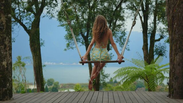 Elegant Long Haired Caucasian Girl Sitting, Posing on the Swing Outside with the Beautiful View on the Bali. Young Lady Enjoying the nature Outdoors with Swinging. Traveling and Tourism Concept.