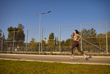 Slim woman jogging alone on a sunny summer morning. Young fit female athlete in sportswear running along a tall wire fence on a city street. Sport, exercise, fitness, outdoor workout concept