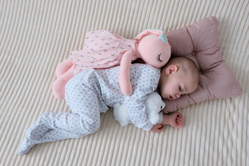 Baby with pink bunny soft toy. A girl of European appearance, wearing a light light jumpsuit, is...
