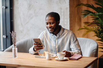 Young smiling African American male manager looking at smartphone screen and reading online news...