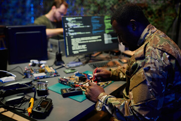 Young serious African American male security officer in camouflage uniform repairing circuit board...