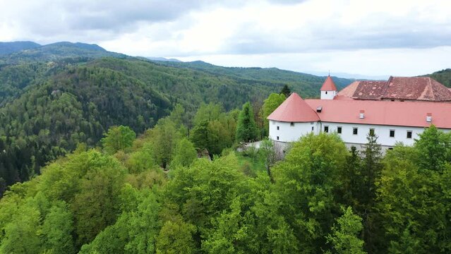 Flying towards a secluded medieval Turjak castle in highland nestled among lush trees and rolling hills, with the clouds, creating a tranquil escape into nature.