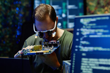 Young military officer or repairman wearing eyewear with small lamps checking details of processor...
