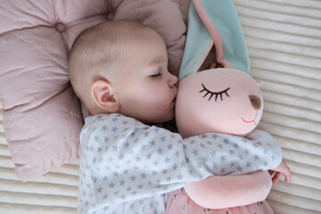 Baby with pink bunny soft toy. A girl of European appearance, wearing a light light jumpsuit, is sleeping in the crib. Sleep schedule, care and hygiene of a child up to one year old.