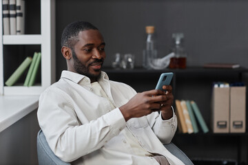 Portrait of adult African American man using smartphone relaxing in chair and scrolling social...