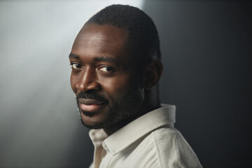 Fototapeta premium Dramatic closeup portrait of African American adult man smiling at camera over shoulder on black background, copy space