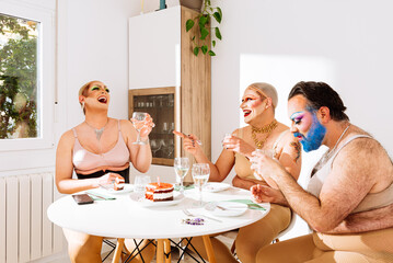 Group of cheerful adult men drag queens in makeup bras sitting at table and laughing