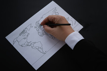 A hand draws a world map with a pencil on a sheet of paper against a dark background. New world...
