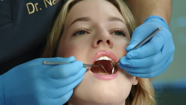 Unrecognizable male dental expert in medical gloves with tooth scraper and concave mirror checking teeth condition of pretty woman with mouth wide open. High quality 4k footage