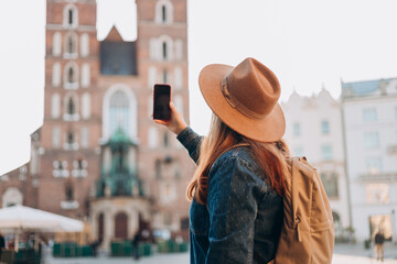 Happy young European tourist with backpack in hat makes photo or video on smartphone on Market Square in Krakow. Traveling Europe in summer. St. Marys Basilica. Vacation concept - obrazy, fototapety, plakaty