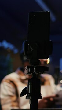 Vertical video Focus on smartphone on tripod recording vlogger show host in blurry background interviewing guest in neon lights studio. Presenter records online podcast episode with mobile phone