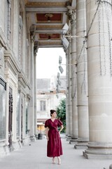 Fototapeta premium Attractive girl with hairstyle, in red dress is walking on street on old building background