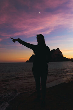 Silhouette of a woman pointing to the left with a pink and orange sunset in the background in Calpe, Spain, vertical image