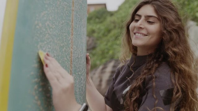 Mulher jovem sorridente na praia aplicando parafina em sua prancha de surf. Cinematico 4k.