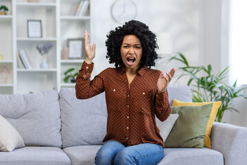 Portrait of a young angry African American woman sitting on the sofa at home, screaming and...