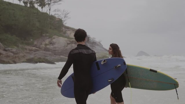 Couple of surfers walking along the beach with their boards under their arms, talking. Rear view. Cinematic 4k.