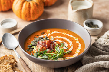 Pumpkin cream soup decorated with cream, pumpkin seeds, bacon, microgreens and olive oil on brown...
