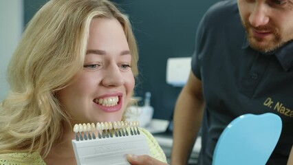 Close up of attractive young woman showing teeth, choosing dentures while male dental expert offers samples of dental implants in modern dentist office. High quality 4k footage