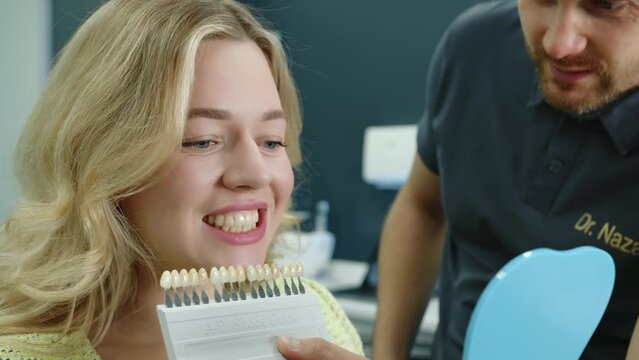 Close up of attractive young woman showing teeth, choosing dentures while male dental expert offers samples of dental implants in modern dentist office. High quality 4k footage