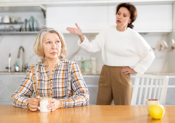 Upset old woman sitting at the kitchen table with her back to middle-aged woman quarreling angrily...