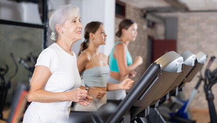 Mature positive woman running on treadmill in modern fitness club