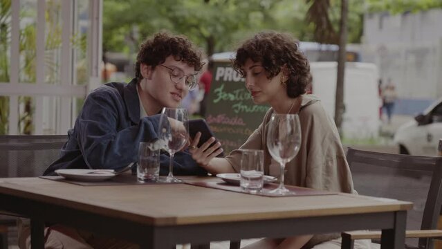 Jovem casal sentado a mesa de um restaurante ao ar livre conversando, usando smartphone. Cinematico 4k.