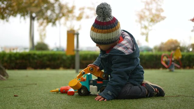 Little boy playing with truck outside at park during autumn day. Child wearing beanie and jacket immersed in play with object