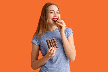 Beautiful young woman eating chocolate bar on orange background