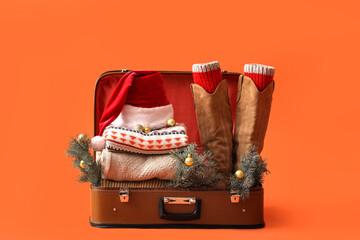 Suitcase with Santa hat, warm clothes, Christmas balls and fir branches on orange background....