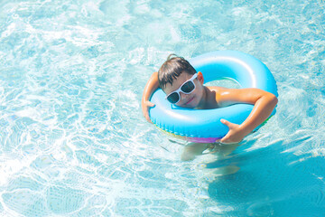 Cute baby boy in white sunglasses swimming with a blue inflatable ring in the clear water of the pool	 - 695623577