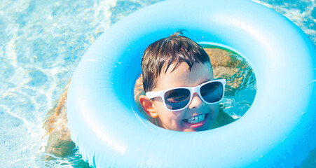 Cute baby boy in white sunglasses swimming with a blue inflatable ring in the clear water of the pool	 - 695623596